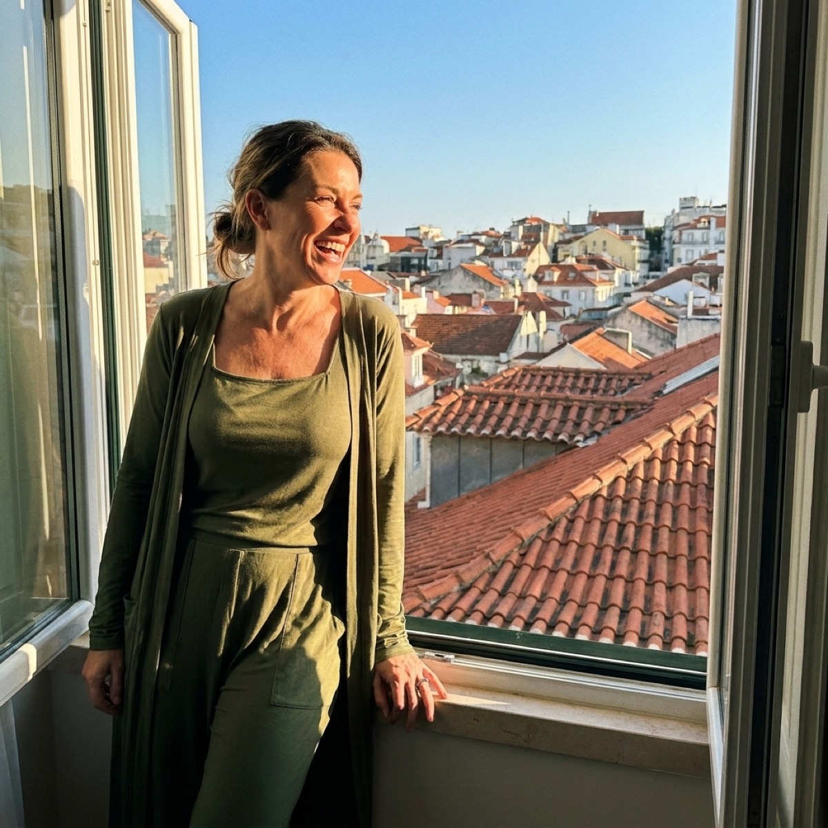 A woman in the olive Essential Set laughing at a hotel window with terracotta rooftops behind her